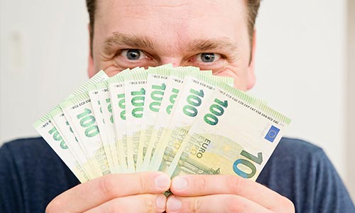 A close-up photo of a smiling man’s face and hands holding fanned out real money bills on a light background