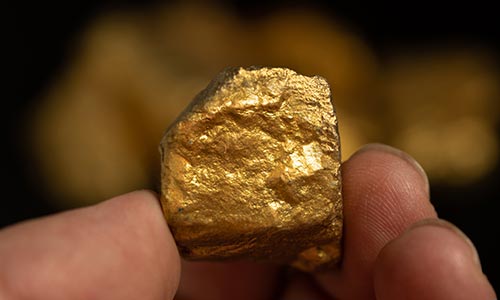 A close-up image of a hand holding a gold nugget against a dark and blurry background